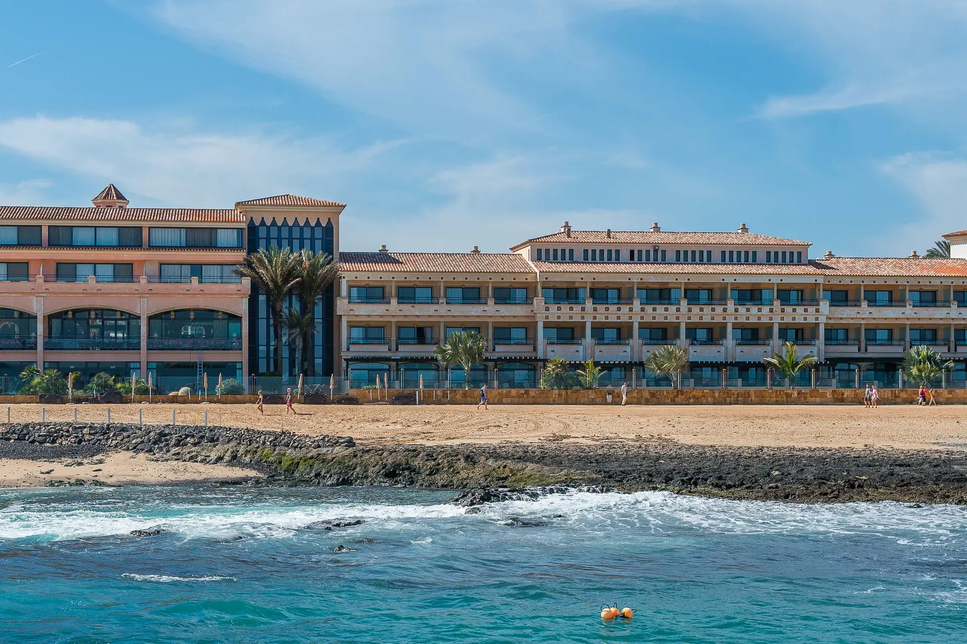 A crescent of golden sand fronted by a beachfront hotel at Playa de la Barreta, Corralejo, with black volcanic rock along the waterline and turquoise water