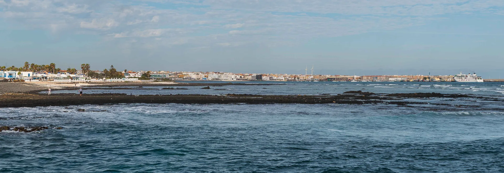A wide view along the rocky Corralejo coastline towards the town and harbour, with wind turbines visible inland