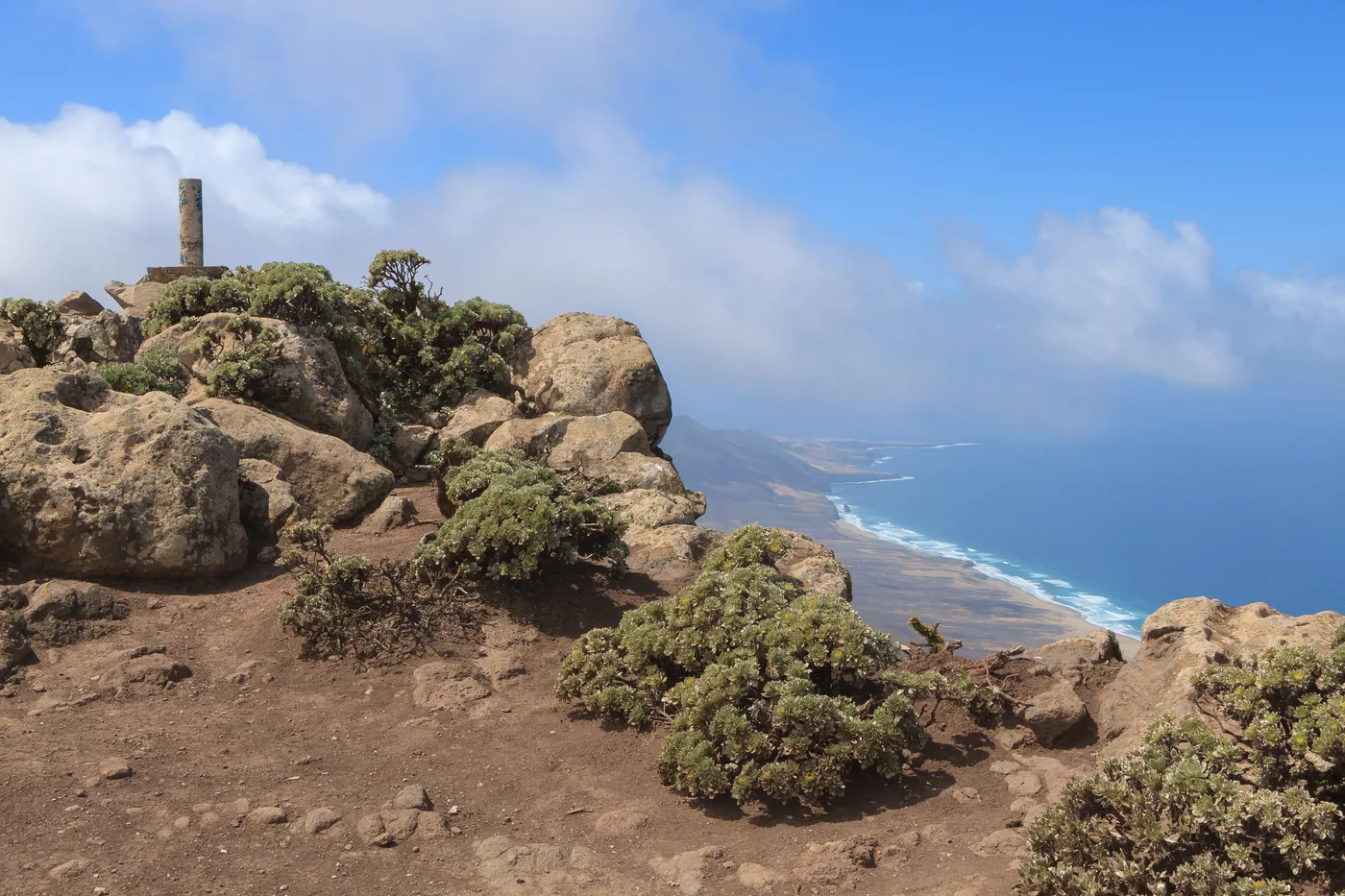 La cima rocosa y batida por el viento del Pico de la Zarza, marcada por un antiguo poste entre rocas y matorral bajo, con una amplia vista sobre la costa de Jandía hacia una playa lejana y el mar
