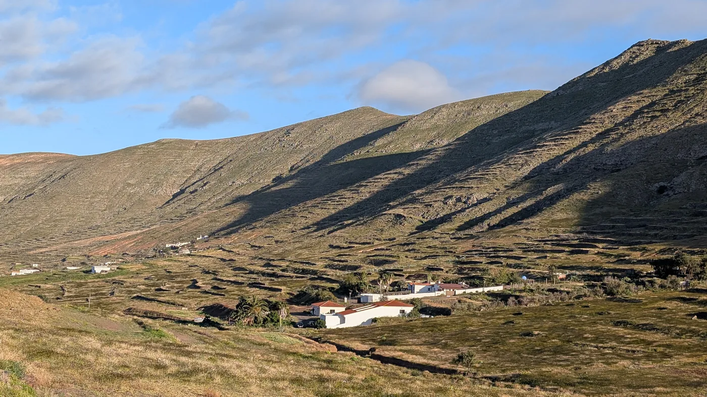 The green, terraced Vallebrón valley seen from above, with whitewashed farmhouses scattered among fields and hills rising on either side under a golden evening light